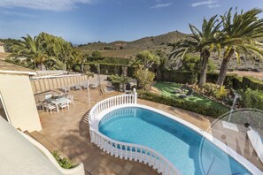 Une piscine en forme de courbe, entourée d’une clôture blanche, avec un espace salon extérieur et une vue sur des collines et des palmiers.