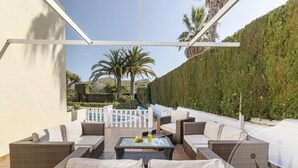 Une terrasse ensoleillée, meublée de meubles en osier, avec une table en verre, et offrant une vue sur une piscine et une végétation luxuriante.