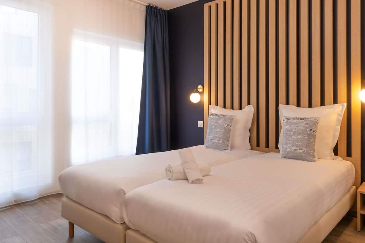 A hotel room with a large bed, two bedside tables, a wooden headboard, and a window with curtains.