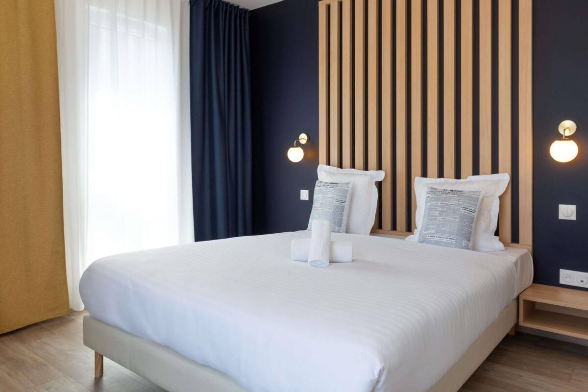 A modern hotel room with a large bed, wooden headboard, and two wall-mounted lights.