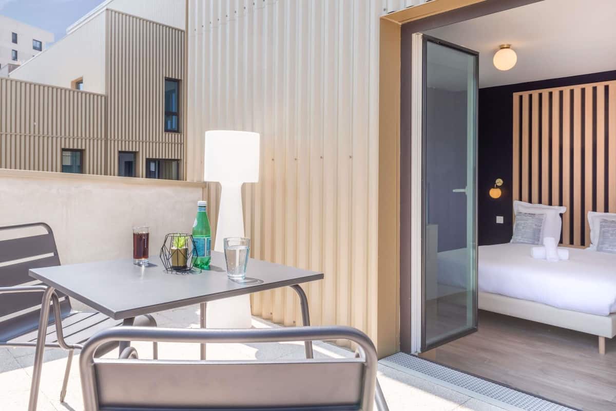 A modern hotel room with a glass door leading to a balcony, a small table with a chair, and a bed with white bedding.