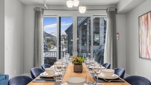 A dining area with a wooden table set for six, a potted plant, and a view of mountains through large windows. - M3- Serene Canmore Retreat- A/C, Free EV Charge, 5BD, 4.5BT, Hot Tub, Mtn View (Canmore)