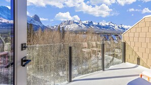 A room with a view of snow-covered mountains, a balcony with a glass railing, and a wooden chair. - M3- Serene Canmore Retreat- A/C, Free EV Charge, 5BD, 4.5BT, Hot Tub, Mtn View (Canmore)