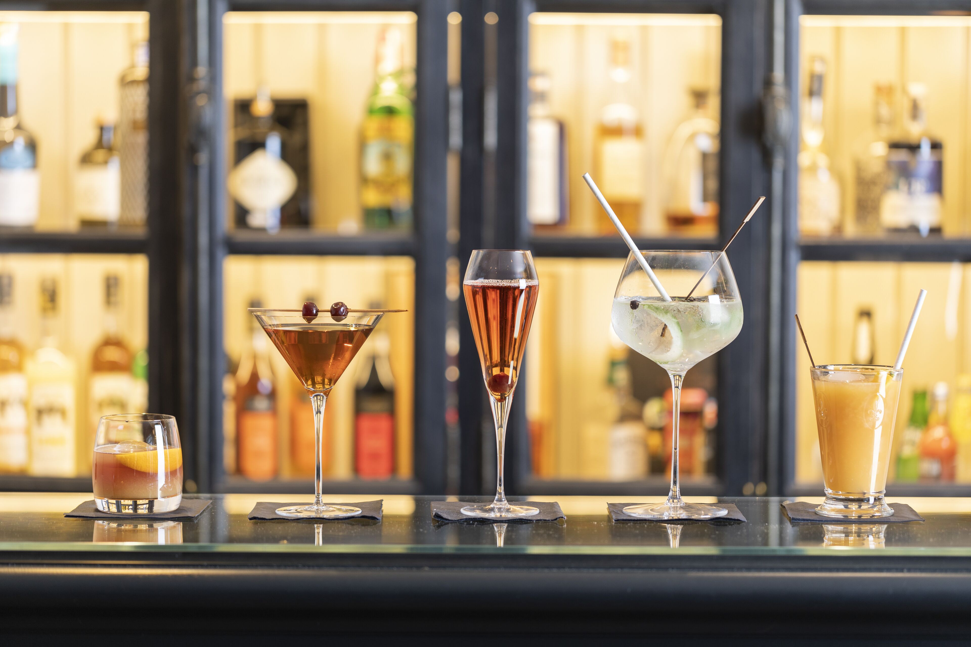 A bar with various drinks, including cocktails and a non-alcoholic beverage, with a background of shelves stocked with bottles.