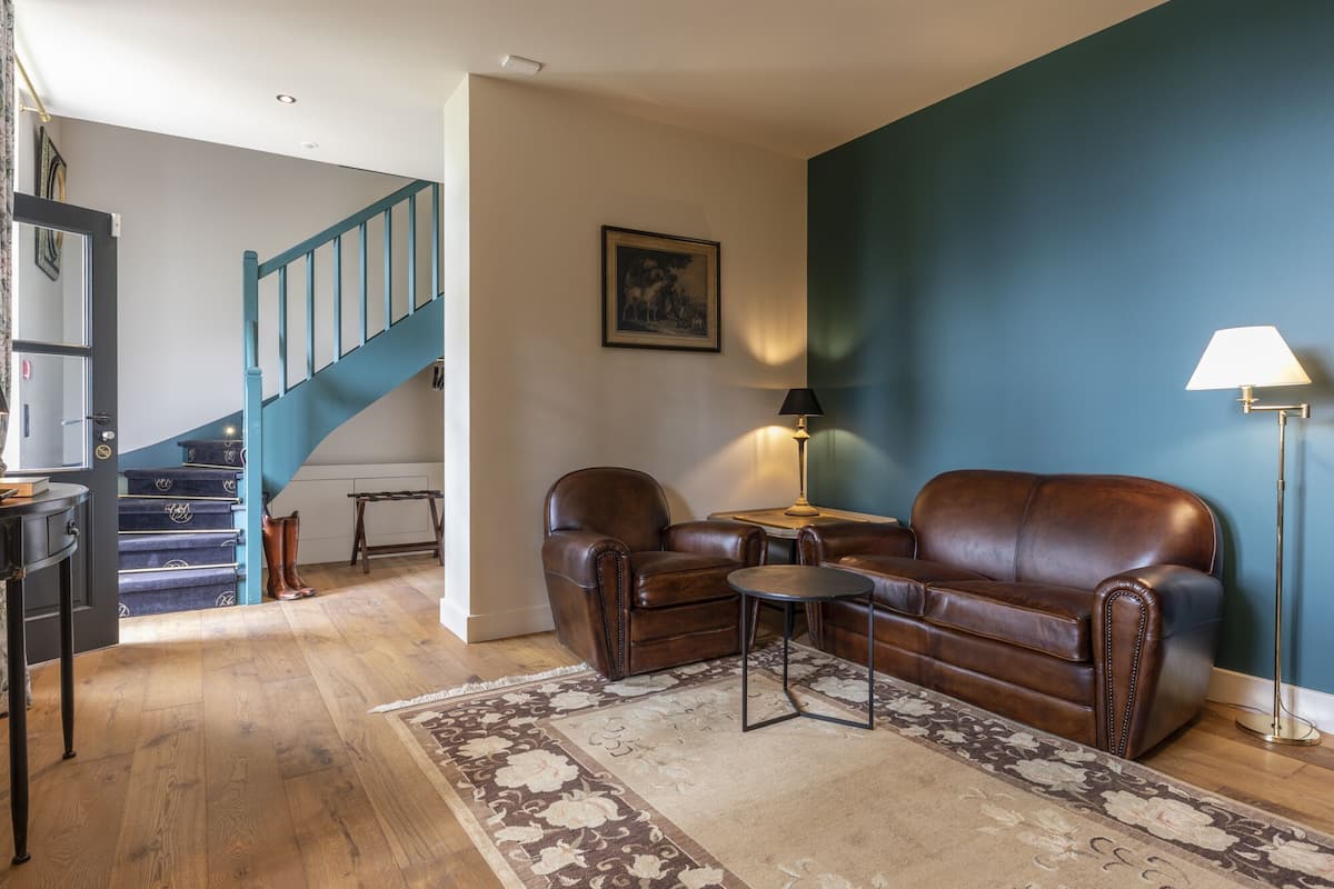 Una sala de estar con un sofá de cuero marrón, una mesita redonda pequeña, una lámpara, una escalera con barandillas verde azulado y una pared de acento azul.