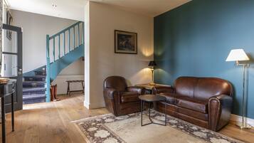 Una sala de estar con un sofá de cuero marrón, una mesita redonda pequeña, una lámpara, una escalera con barandillas verde azulado y una pared de acento azul.
