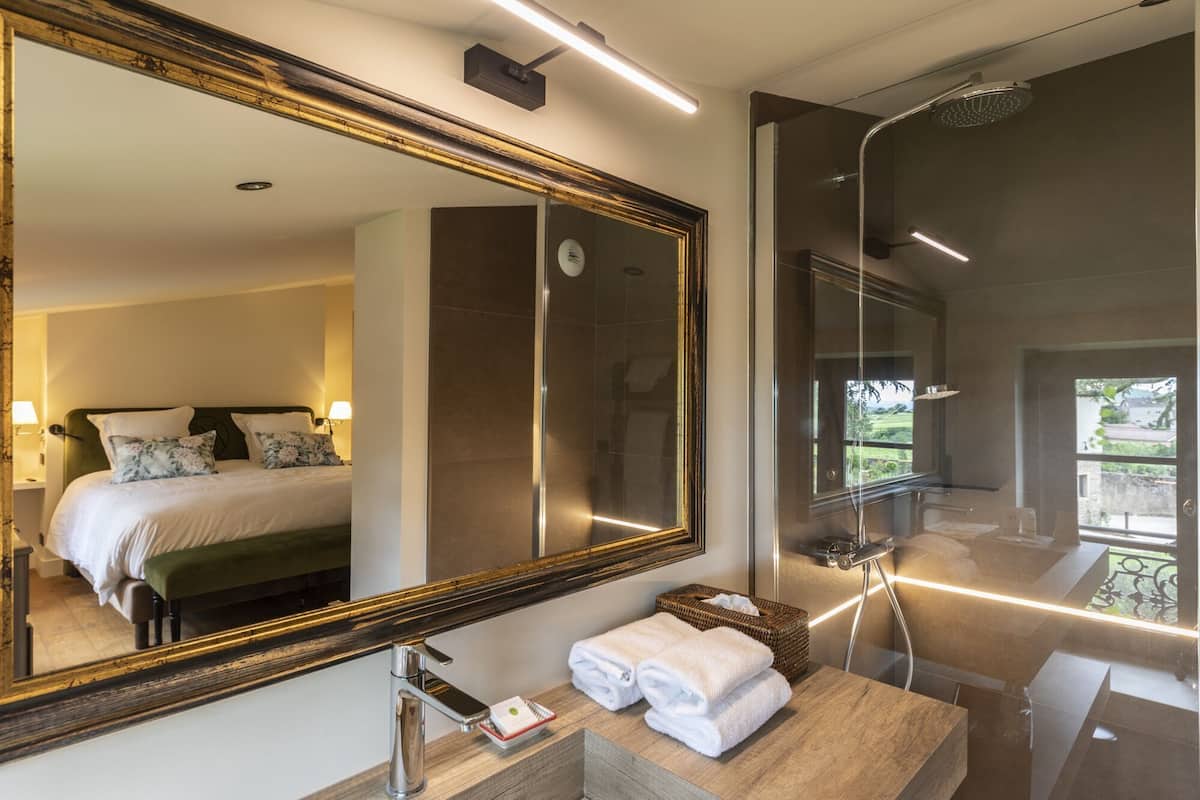 Un baño moderno con un espejo grande, una ducha y una cama visible en el reflejo.