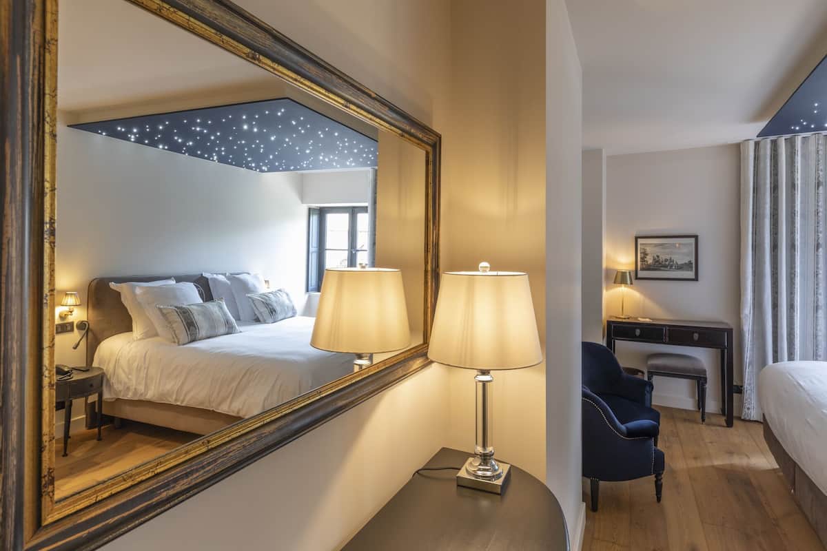 Habitación de hotel con un espejo grande, dos lámparas de noche, una cama con almohadas, un escritorio con una silla y una mesita de noche con lámpara.