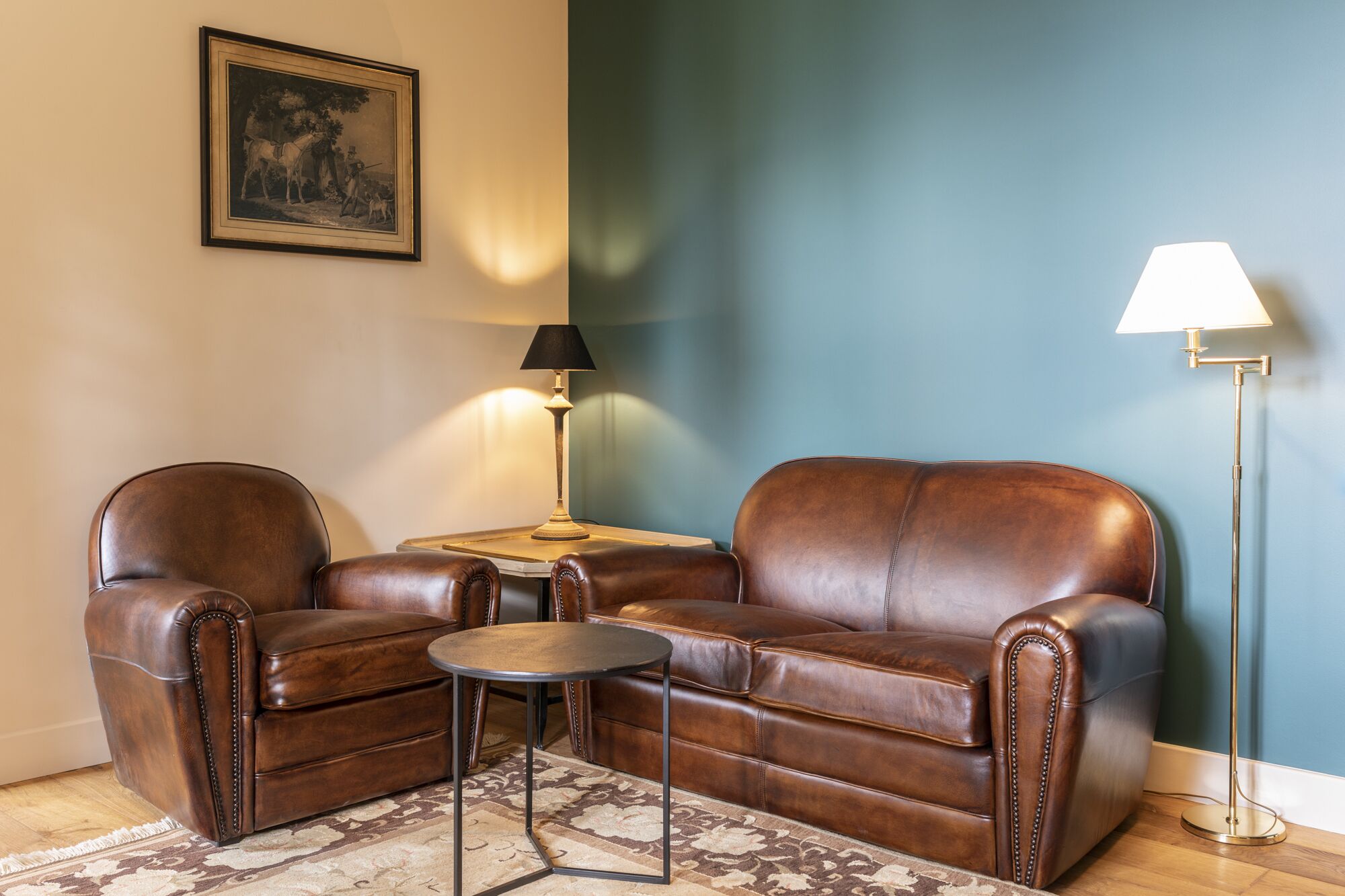 A living room with a brown leather sofa, a small round table, a lamp, and a framed picture on the wall.