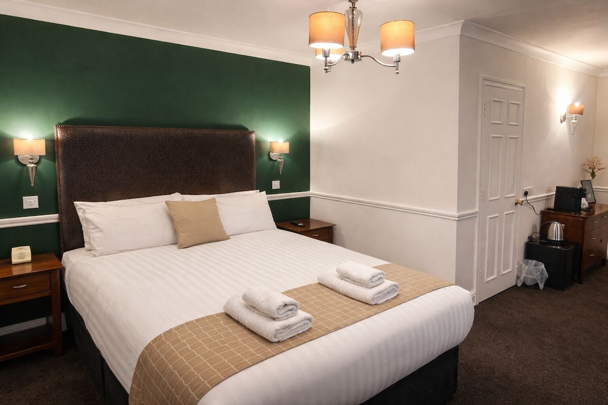 A neatly made bed with white linens and a brown throw, a nightstand with a lamp, and a wall-mounted light fixture.