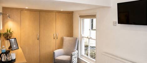 A room with a wooden wardrobe, a grey armchair, a coffee station, a radiator, and a TV on the wall.