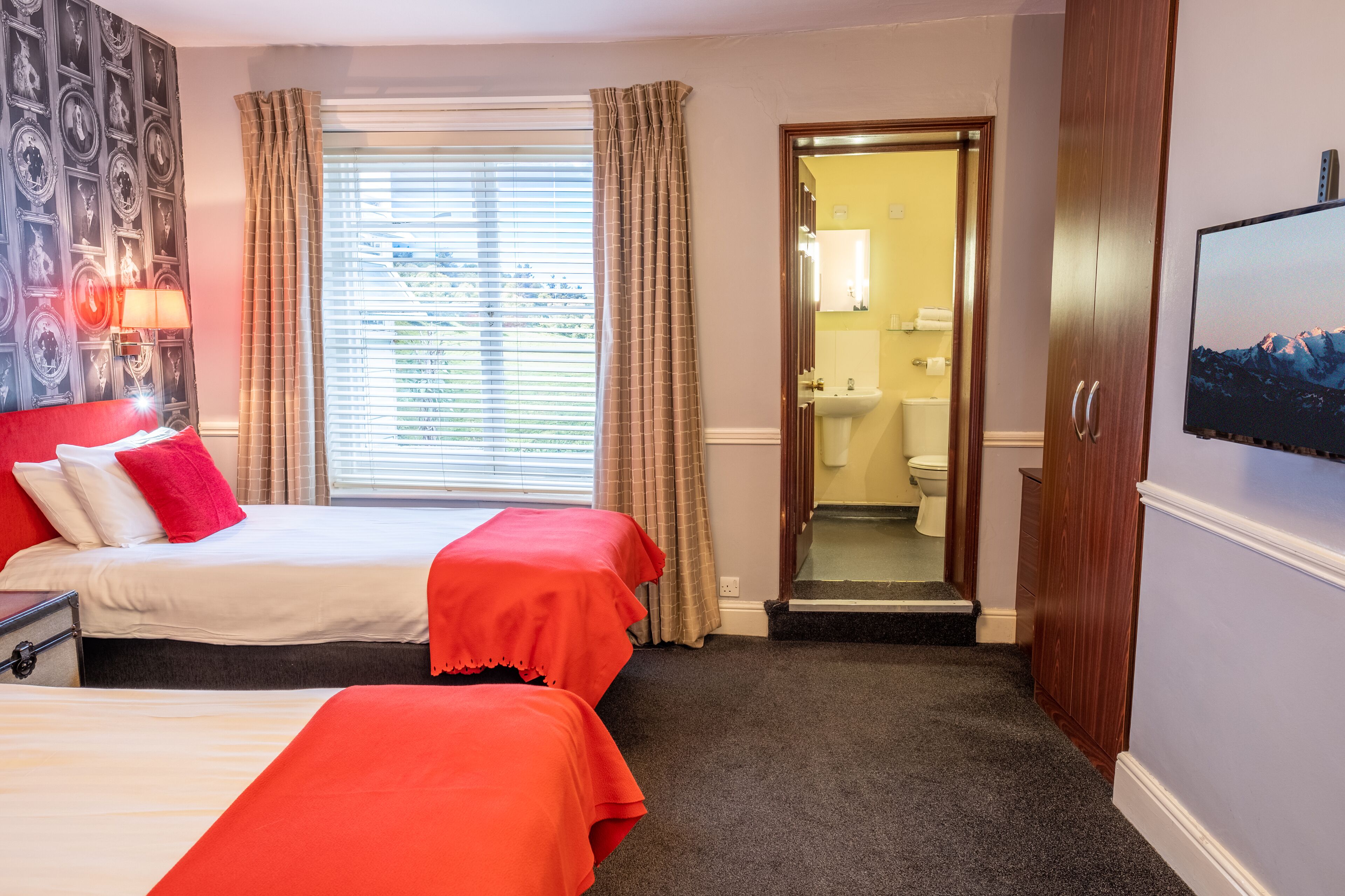 A hotel room with two beds, a large window with curtains, a bathroom visible through an open door, and a television on the wall.