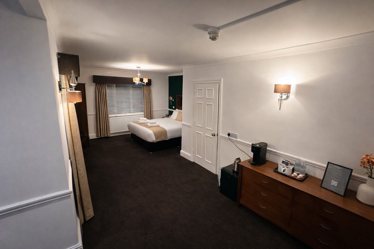 A hotel room with a bed, a dresser, a desk with a coffee maker, and a wall-mounted lamp.
