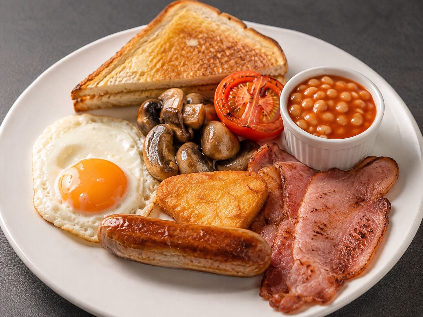 A plate of breakfast food including toast, eggs, mushrooms, sausages, bacon, beans, and tomato.