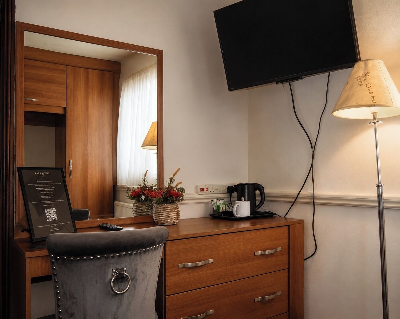 Quarto de hotel com escrivaninha de madeira, uma cadeira, televisão de tela plana na parede, luminária e cafeteira.