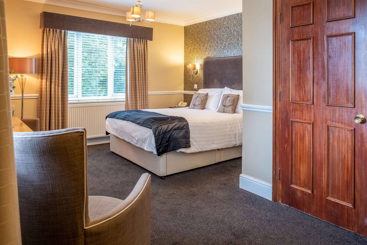 A hotel room with a bed, a chair, a window with curtains, a wall-mounted lamp, and a wooden door.