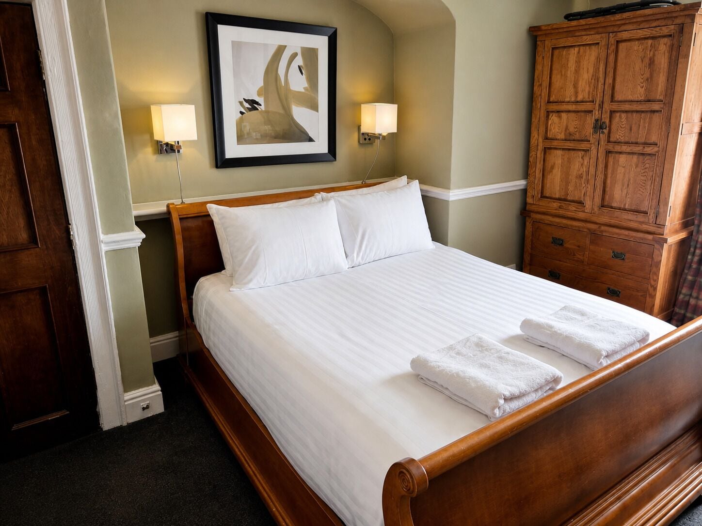 A neatly made bed with white linens and two pillows, a wooden headboard, a framed artwork on the wall, and a wooden wardrobe with drawers.