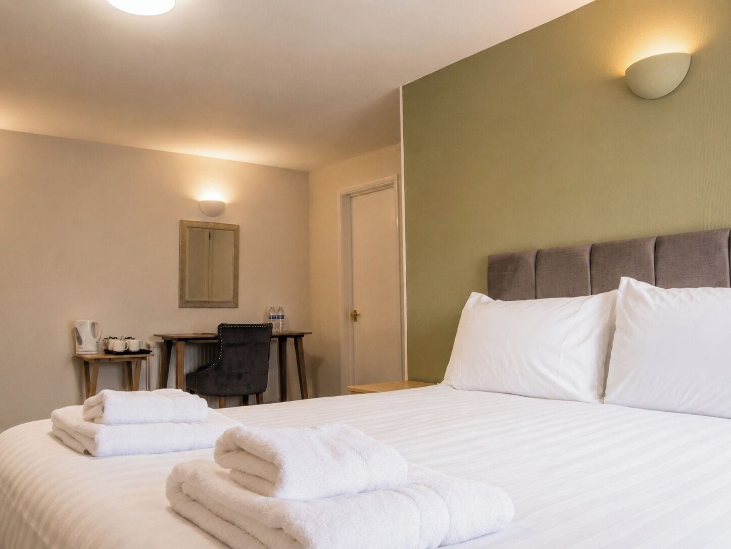A hotel room with a bed, two towels, a desk with a chair, a mirror, and a wall-mounted light fixture.