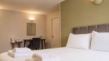 A hotel room with a bed, two towels, a desk with a chair, a mirror, and a wall-mounted light fixture.