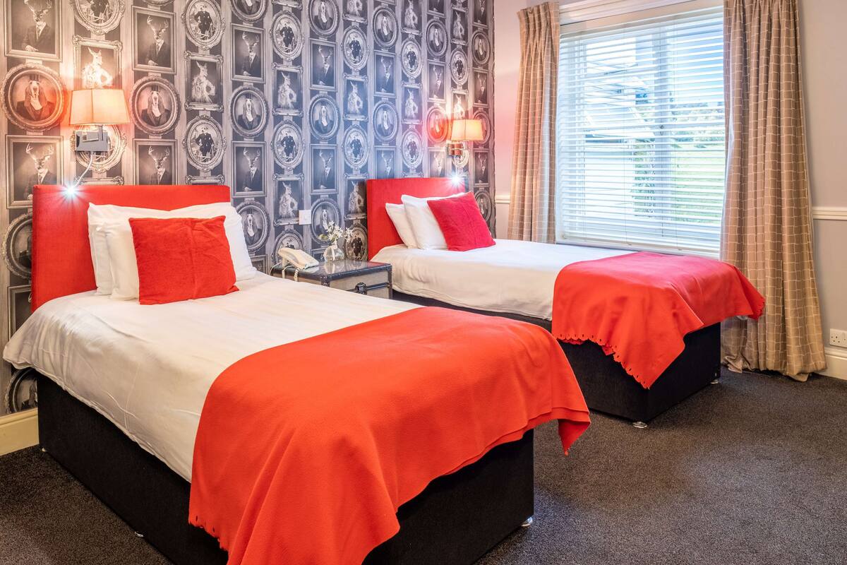 Two beds with red bedding, a patterned wallpaper, and a window with curtains.