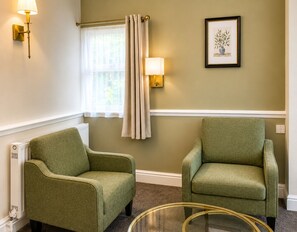 Ein Zimmer mit zwei Sesseln, einem fensterbelaubten Fenster, Wandlampen und einem gerahmten Bild.