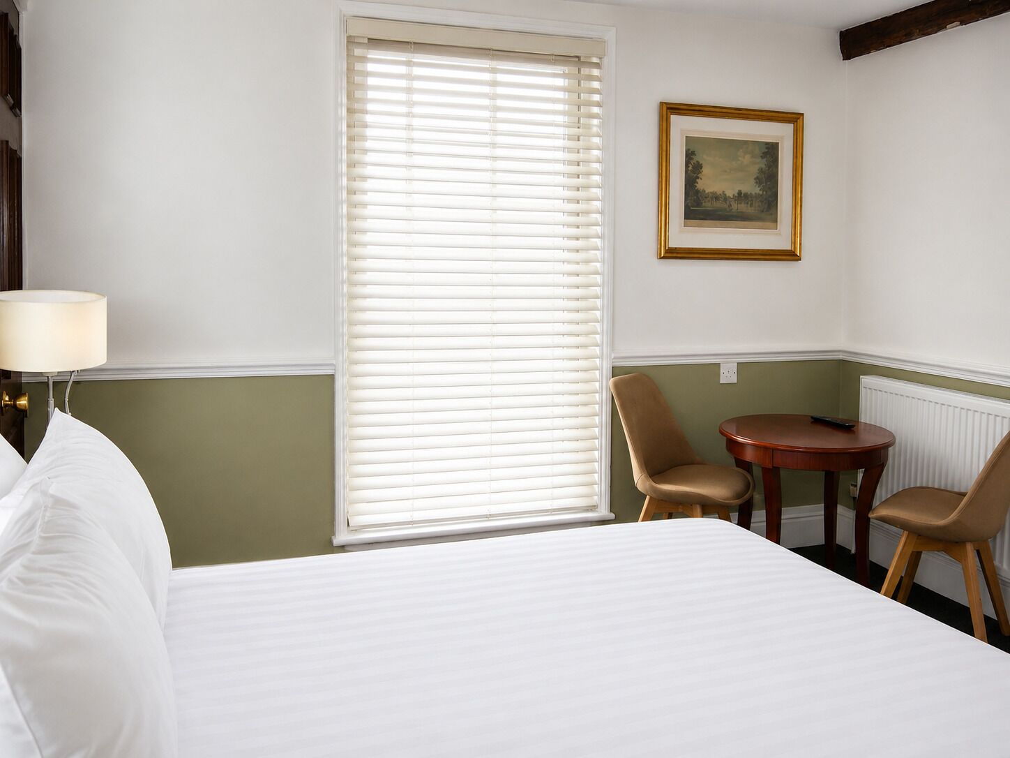 Una habitación de hotel con una cama, una mesa, dos sillas, una lámpara y un cuadro en la pared.