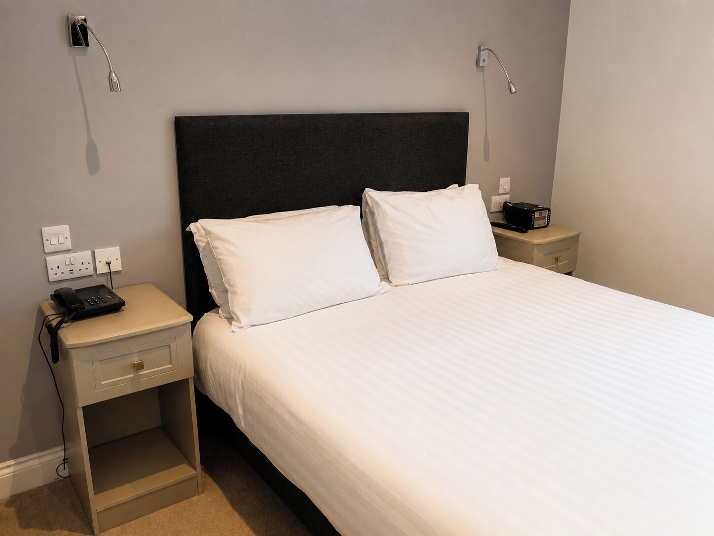 A neatly made bed with white linens, a bedside table with a phone and a radio, and a nightstand with a lamp.