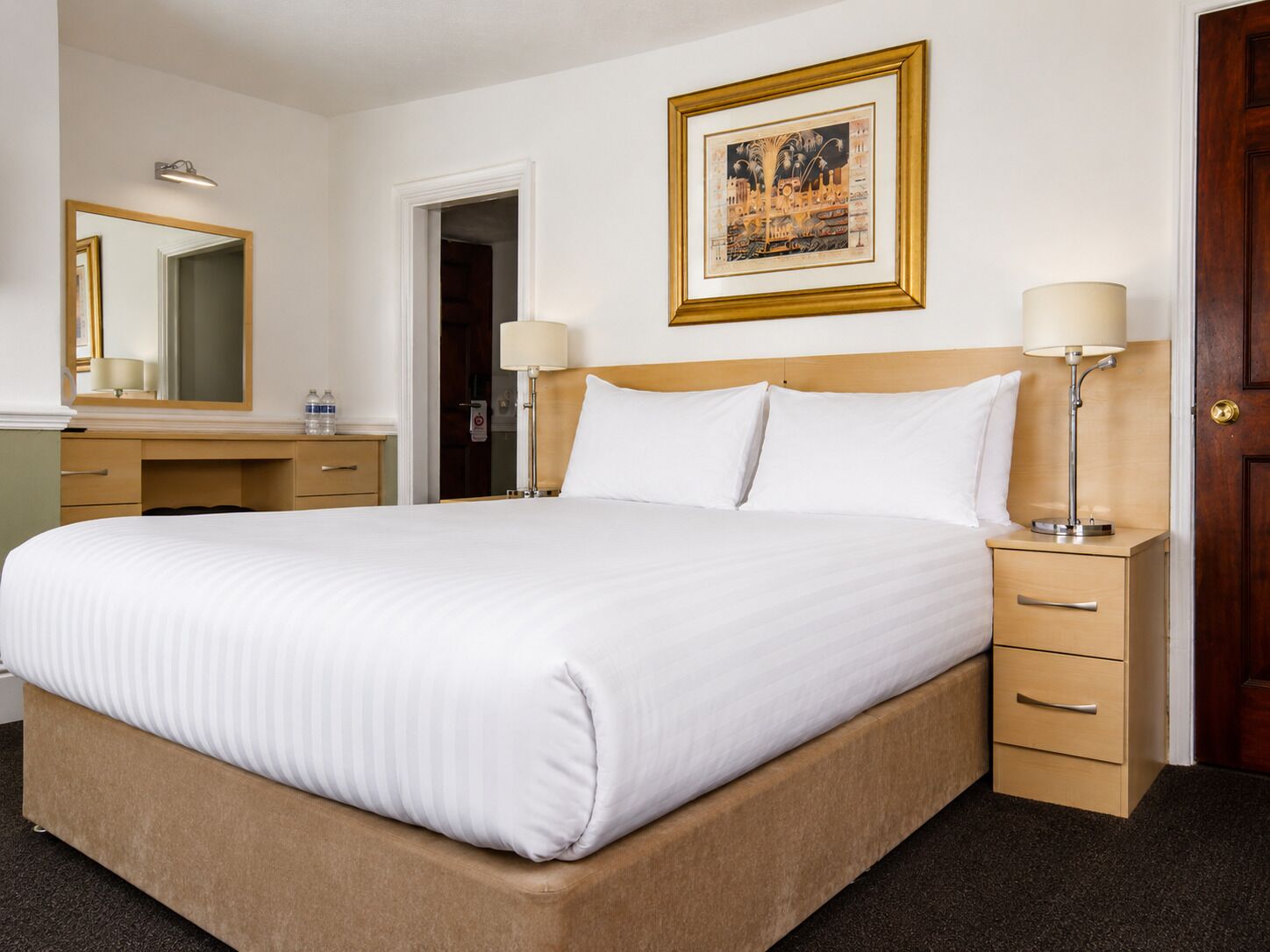 Ein Hotelzimmer mit einem großen Bett, Nachttischen, einem Schreibtisch, einem Spiegel und einem gerahmten Bild an der Wand.