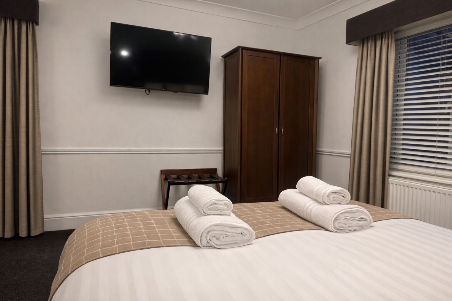 A hotel room with a bed, a TV mounted on the wall, a wardrobe, and a window with blinds.