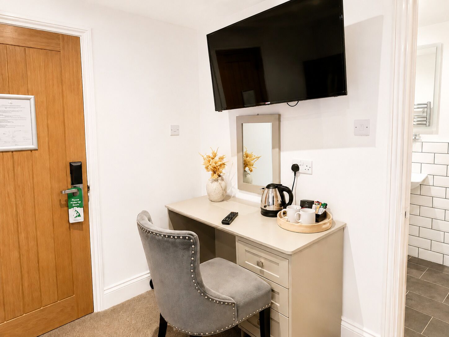 A hotel room with a desk, a chair, a television mounted on the wall, a mirror, and a small table with a kettle and cups.
