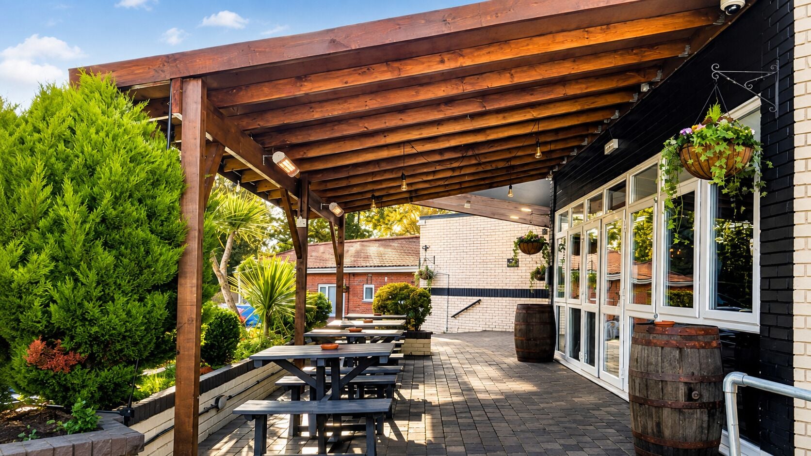 Een terras buiten met houten tafels en banken, een bakstenen muur en hangende plantenbakken.