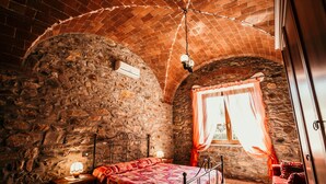 A bedroom with a stone wall, a bed with a red quilt, a nightstand with a lamp, and a window with orange curtains.