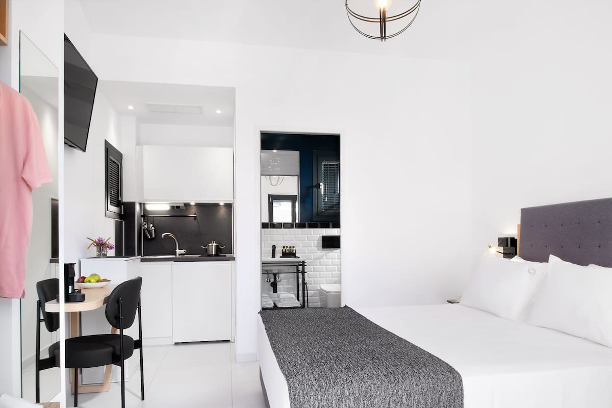 Ett modernt hotellrum med en säng, ett skrivbord med en stol, ett litet pentry och ett badrum som syns genom en öppen dörr.
