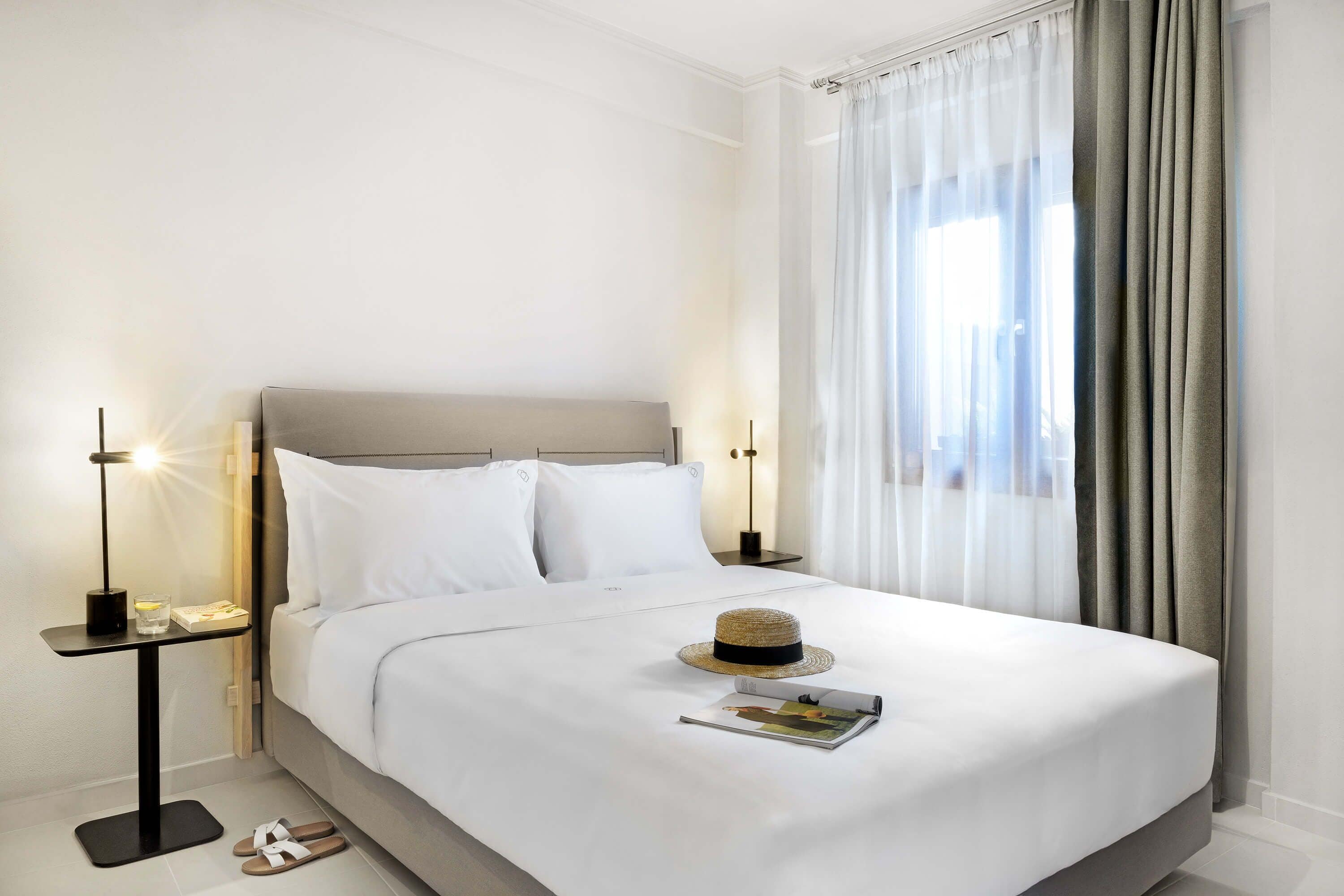 A neatly made bed with white linens, a bedside table with a lamp, and a straw hat on the bed.