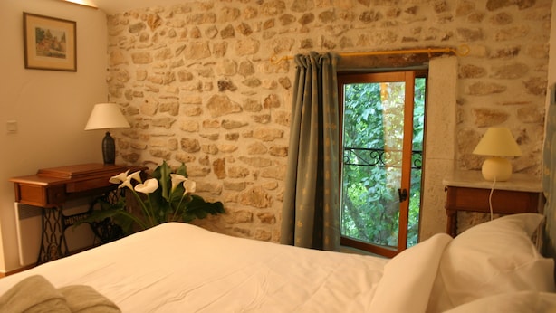 Quarto com parede de pedra, cama com roupa de cama branca, escrivaninha com abajur e janela com cortinas.