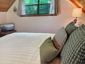 A bedroom with a bed, pillows, a TV on a wooden stand, a window with curtains, and a lamp on a bedside table.