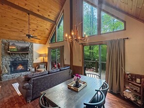 Un interior acogedor con chimenea de piedra, techo de madera, zona de comedor con mesa y sillas, y ventanas grandes que ofrecen vistas al exterior.