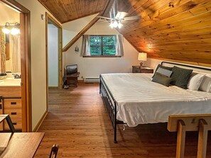 A bedroom with a wooden ceiling, a bed with pillows, a nightstand, a window with a view of greenery, and a television on a stand.