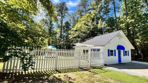 A small white building with a blue door and windows, surrounded by a white picket fence and trees. - Santorini Cave Style Cottage with Fenced-in Yard (Chapel Hill)
