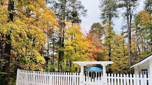 A white picket fence surrounding a gazebo with a blue roof, set against a backdrop of autumn-colored trees. - Santorini Cave Style Cottage with Fenced-in Yard (Chapel Hill)