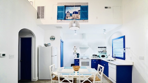 A compact kitchen with a dining area, featuring a central island, white cabinets, and blue accents. - Santorini Cave Style Cottage with Fenced-in Yard (Chapel Hill)
