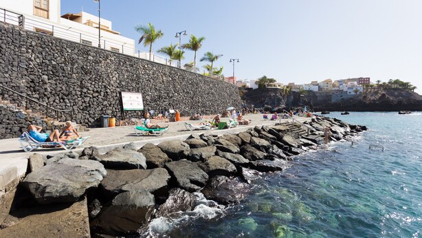 A coastal area with a stone wall, beach chairs, and people relaxing by the water.