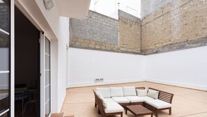 A spacious patio with a sectional sofa and a wooden table, a sliding glass door leading to an indoor area, and a brick wall.