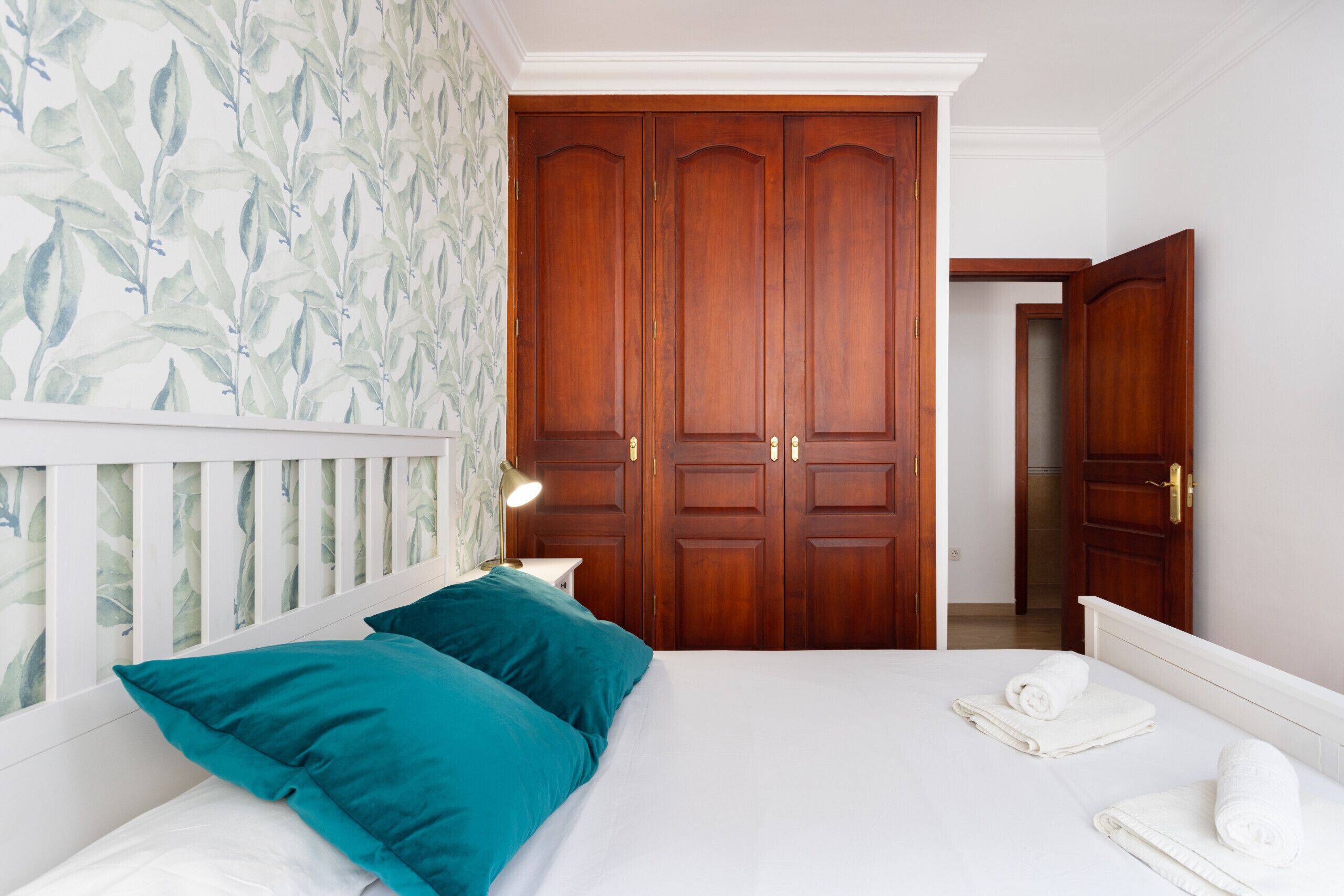 A bedroom with a bed, teal pillows, a nightstand with a lamp, and a wooden wardrobe.