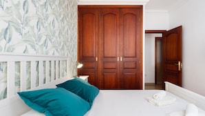 A bedroom with a bed, teal pillows, a nightstand with a lamp, and a wooden wardrobe.