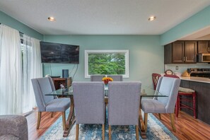 A dining area with a glass table, grey upholstered chairs, a blue patterned rug, a TV on the wall, and a view of greenery outside. - Bent Creek 28 at Rumbling Bald (Lake Lure)