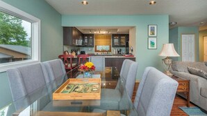 Un coin repas avec une table en verre, des chaises à revêtement gris et une petite table d’appoint en bois.