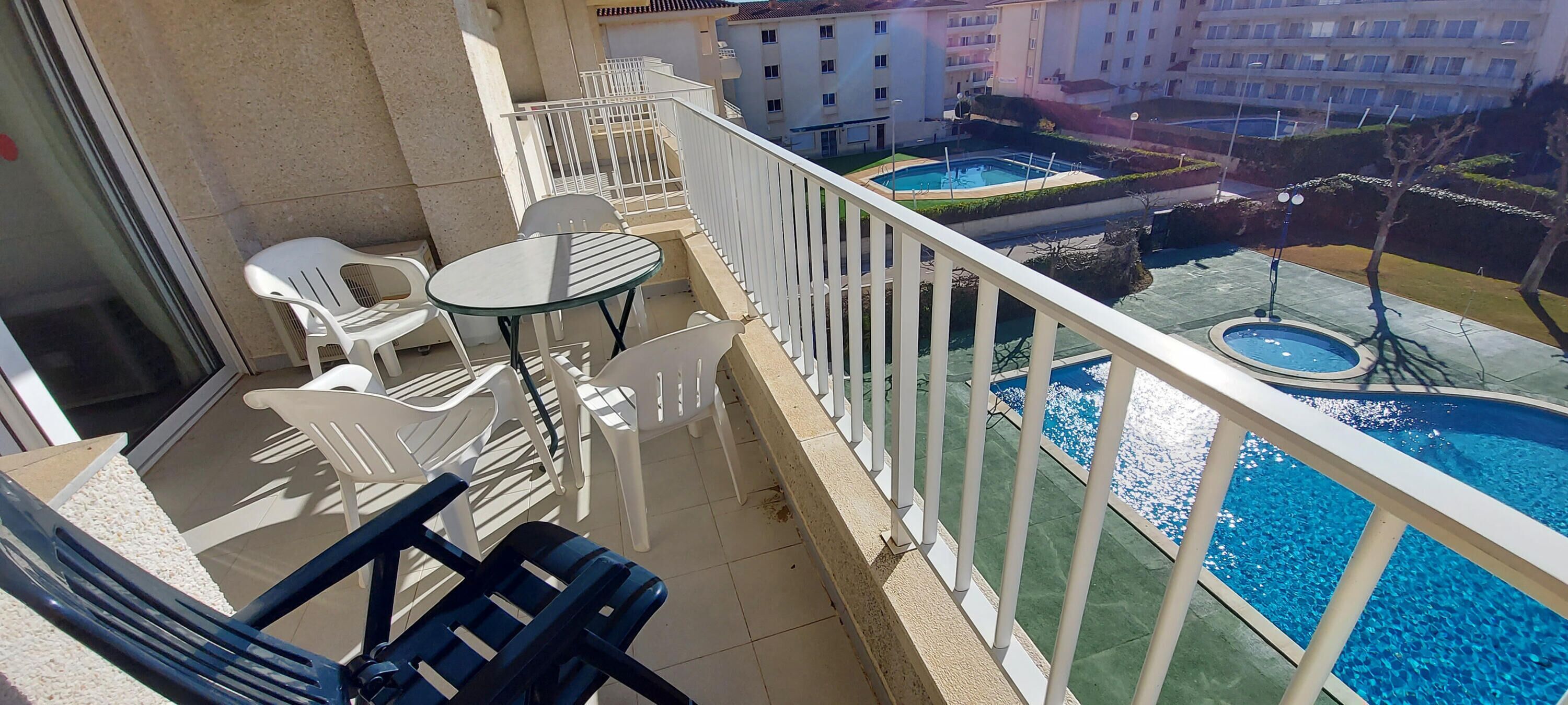Ein Balkon mit weißen Plastikstühlen und einem Glastisch, der einen Swimmingpool und einen angelegten Garten mit einer Fontäne blickt.