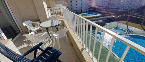 Ein Balkon mit weißen Plastikstühlen und einem Glastisch, der einen Swimmingpool und einen angelegten Garten mit einer Fontäne blickt.