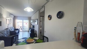 Ein modernes Wohnzimmer mit einer Couch, einem Esstisch und Stühlen. Ein Fernseher steht auf einem Schrank, eine Deckenleuchte ist angebracht, und eine Tür führt in einen weiteren Raum.